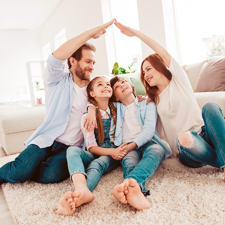 Happy family with two kids sitting on carpet, mom and dad making roof figure with hands arms over heads. New building residential house purchase apartment concept. 